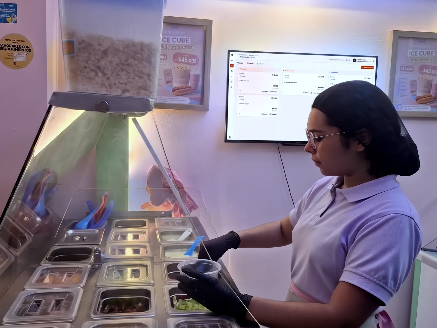 Una empleada de Ice Cube preparando un pedido en la barra de toppings, con el tablero de ventas de Dayvent en la pantalla del fondo.